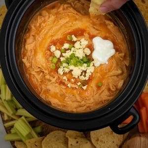 Slow cooker filled with cheesy buffalo chicken dip surrounded by festive holiday decor